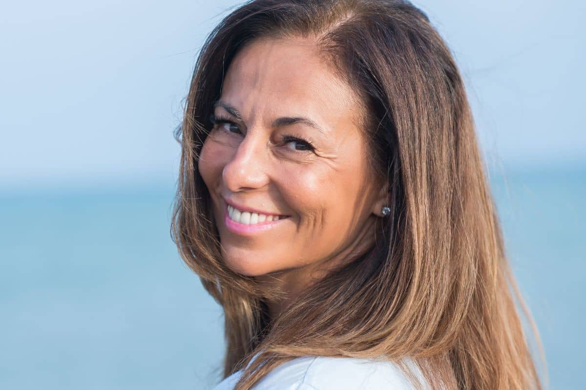 Smiling middle-aged woman with healthy teeth enjoying a sunny day by the ocean, representing dental implant longevity.