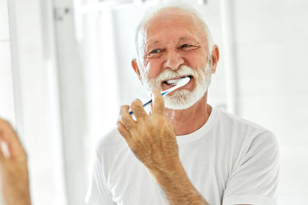 Smiling older man brushing his teeth in a bright bathroom, demonstrating daily care to maintain dental implants.