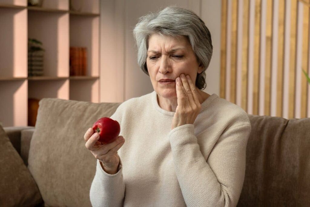 Concerned older woman holding an apple and touching her jaw, indicating possible dental implant or crown issue.